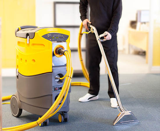 Man in black with white sneakers dry vacuuming a grey floor with a yellow and brown dry vac with a silver nozzle and broom